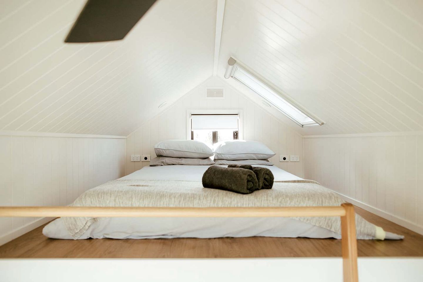 A small, white, attic-style bedroom with a sloped ceiling, a single bed with pillows, a wooden headboard, and a skylight.