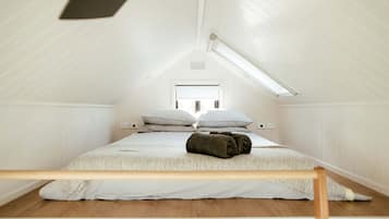 A small, white, attic-style bedroom with a sloped ceiling, a single bed with pillows, a wooden headboard, and a skylight.