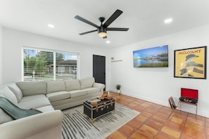 A modern living room with a sofa, a ceiling fan, a red suitcase, and a framed picture on the wall. - Ocean Breeze Cottage: Private Oasis Near Boynton Beach & Delray Beach. (Boynton Beach)