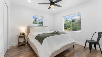 Un dormitorio con una cama, una silla, una mesita de noche, un ventilador de techo y dos ventanas.