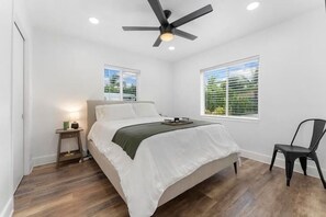 A bedroom with a bed, a chair, a nightstand, a ceiling fan, and two windows.