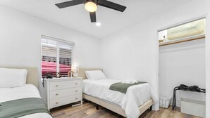 A bedroom with two beds, a nightstand, a window with blinds, a ceiling fan, and a radiator. - Ocean Breeze Cottage: Private Oasis Near Boynton Beach & Delray Beach. (Boynton Beach)