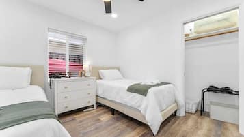 Ein Schlafzimmer mit zwei Betten, einem Nachttisch, einem Fenster mit Jalousien, einem Deckenventilator und einer Heizung.