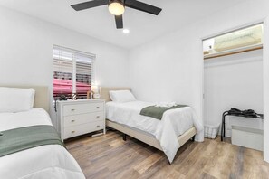 A bedroom with two beds, a nightstand, a window with blinds, a ceiling fan, and a radiator. - Ocean Breeze Cottage: Private Oasis Near Boynton Beach & Delray Beach. (Boynton Beach)