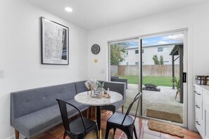 Ein Zimmer mit grauem Sofa, einem runden Tisch und Stühlen, einer Uhr an der Wand und einer Glastür, die in einen Garten führt.