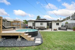 A backyard with a swimming pool, a wooden deck, a covered patio, and a fenced area.