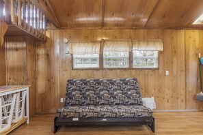 A room with wooden walls and ceiling, a patterned sofa, a small table, and two windows with curtains. - Deluxe Cabin - Our Deluxe Cabin has two rooms (1 bedroom) with a Queen bed, futon or queen sofa, and 4 twin beds in a loft. Complete with a bathroom, kitchen stocked with dishes, toaster, and coffee pot, fireplace, and screened  porch. Non-smoking. M (Gore)