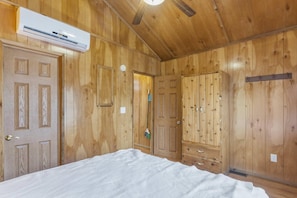 A room with wooden walls, a bed with a white duvet, a ceiling fan, and an air conditioning unit. - Deluxe Cabin - Our Deluxe Cabin has two rooms (1 bedroom) with a Queen bed, futon or queen sofa, and 4 twin beds in a loft. Complete with a bathroom, kitchen stocked with dishes, toaster, and coffee pot, fireplace, and screened  porch. Non-smoking. M (Gore)