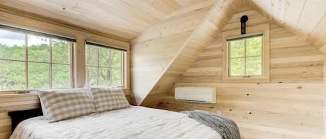Un dormitorio con techo inclinado, dos ventanas, una cama con almohada a cuadros y un equipo de aire acondicionado.