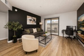 A modern living room with a sectional sofa, a coffee table, a TV, and a balcony with outdoor furniture. - Stylish 2BR in Tempe | Near ASU w/ Pools & Gym (Tempe)
