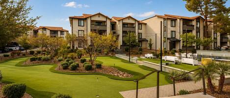 A golf course with a putting green, surrounded by a landscaped area with trees and shrubs, and a building with multiple balconies in the background.
