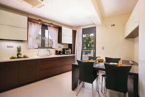 A modern kitchen with a dining area, featuring a large window, dark wooden cabinets, and a breakfast setting with fruits and juice. -     Lamon Villa | Elegant Seaside Escape with Private Pool (Myrthianos Plakias)