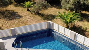 Ein Schwimmbecken mit Fliesen umgeben, umrahmt von Gras und Bäumen. Liegestühle und ein Tisch mit Obstschale und Getränken sind vorhanden.