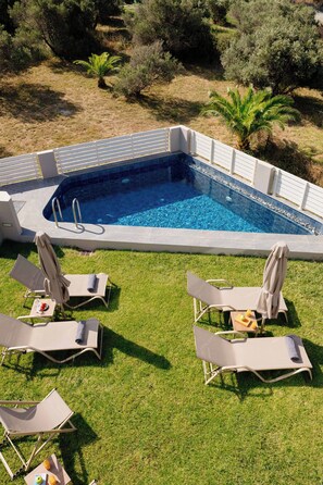 Ein Schwimmbecken mit Fliesen umgeben, umrahmt von Gras und Bäumen. Liegestühle und ein Tisch mit Obstschale und Getränken sind vorhanden.