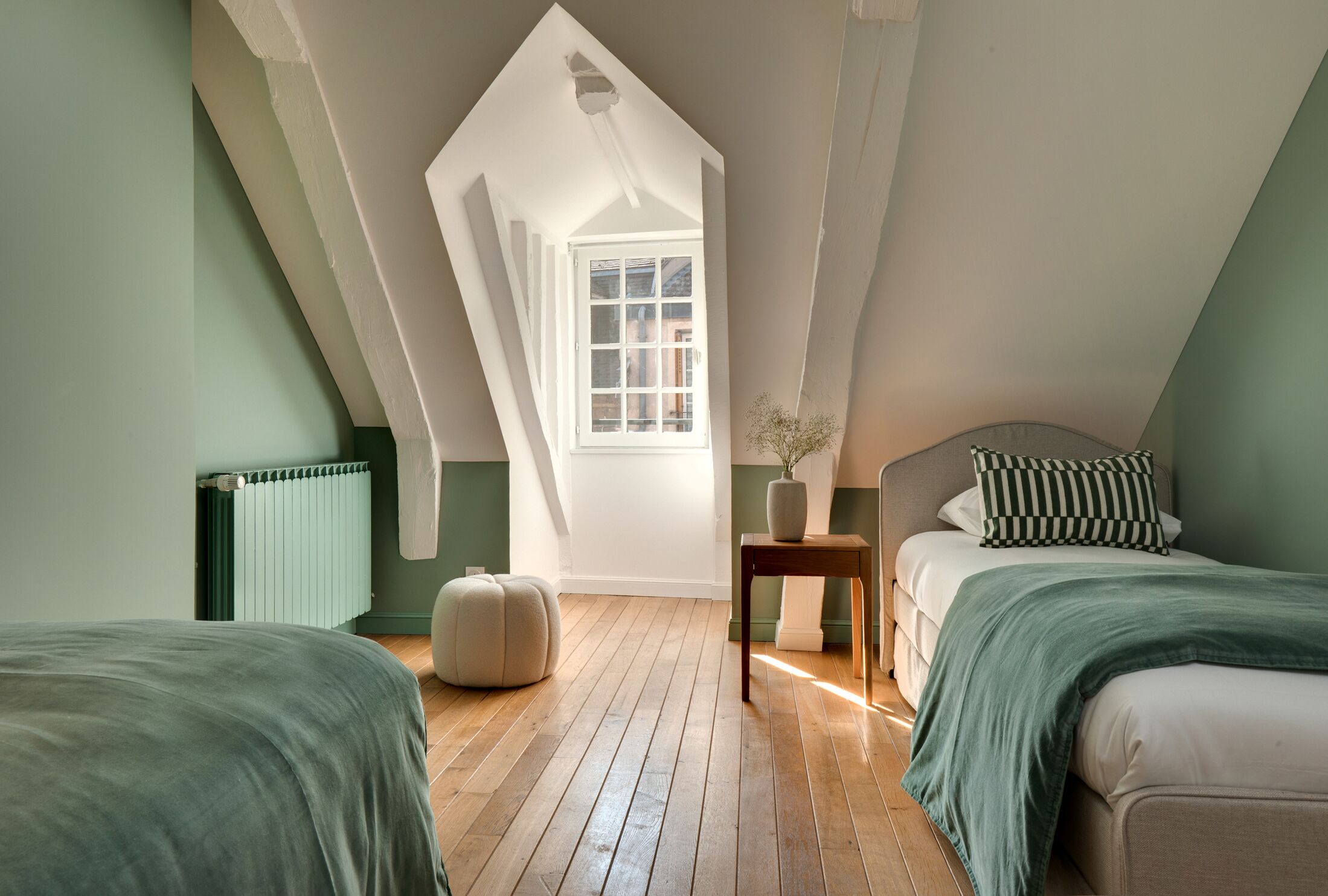 Un dormitorio con techo inclinado, una ventana, un radiador, una mesita de noche con un florero y dos camas con almohadas a rayas.