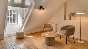 Et moderne loftværelse med synlige bjælker, et stort vindue, et rundt træsofabord, to fletstole, en gulvlampe og en radiator.