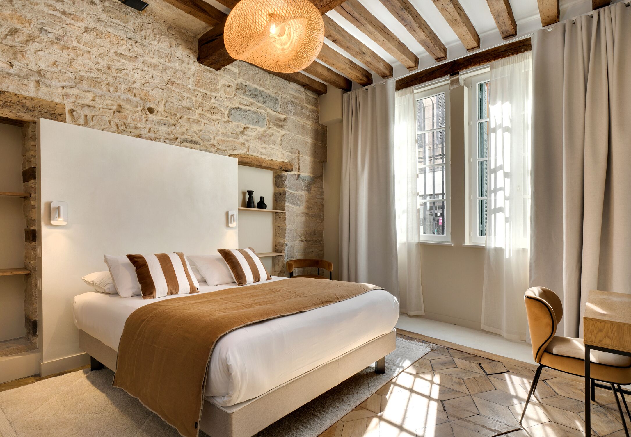 Un dormitorio con pared de piedra, una cama con manta marrón, cabecera de madera, lámpara colgante y una ventana con cortinas.