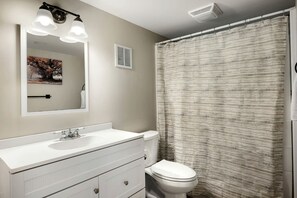 Une salle de bain avec un lavabo blanc, un miroir et des toilettes. On y trouve un rideau de douche et une bouche d’aération au plafond.