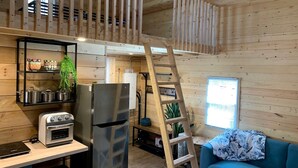 Eine kleine Küche mit Kühlschrank, Herd und Regalen mit Kochgeschirr. Eine Holzleiter steht in der Mitte, und ein blaues Sofa befindet sich im Vordergrund.