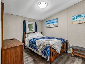 Une chambre à coucher avec un lit en bois, une table de chevet, un banc, une fenêtre avec des rideaux et un tableau représentant une plage accroché au mur.