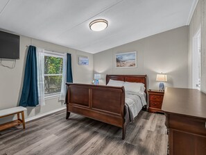 A bedroom with a wooden bed, a bedside table, a desk, a TV mounted on the wall, and a window with curtains.
