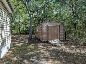 Eine Holzlagerhütte mit Tür und einer kleinen Rampe, umgeben von Bäumen und einem Haus.