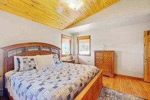 Une chambre à coucher avec une tête de lit en bois, un lit recouvert d’une courtepointe, une table de chevet, une commode et une fenêtre avec des stores.