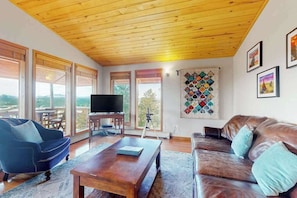 Un salon avec un plafond en bois, un téléviseur, une grande fenêtre et une œuvre d’art murale.