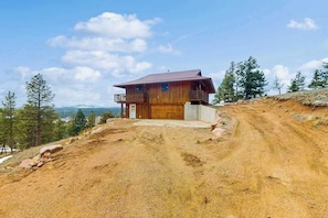 Eine Holzhütte mit Balkon, gelegen an einem Feldweg, umgeben von Bäumen, mit blauem Himmel darüber.