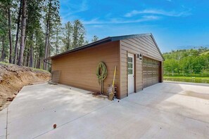 A single-story building with a garage, a concrete driveway, and a landscape with trees and a hill in the background. - Two Bit Cabin - Secluded Cabin • Hot Tub & ATV Trails (Deadwood)