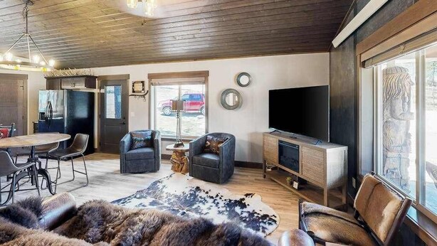 A modern living room with a wooden ceiling, a flat-screen TV, leather sofas, a dining area with a round table, and a large window. - Rost Place Round-Up - Dog-Friendly Cabin • Mtn Views & BBQ (Sturgis)