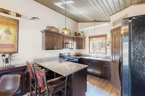 A kitchen with dark wooden cabinets, a granite countertop, stainless steel appliances, and a large refrigerator. - Rost Place Round-Up - Dog-Friendly Cabin • Mtn Views & BBQ (Sturgis)