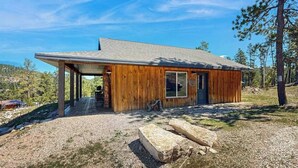 A wooden cabin with a covered porch, a front door, and a window. - Rost Place Round-Up - Dog-Friendly Cabin • Mtn Views & BBQ (Sturgis)
