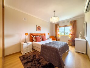 A bedroom with a bed, nightstands, a dresser, a chandelier, a window with curtains, and a city view. - Refúgio do Mar by ACasaDasCasas (Ericeira)