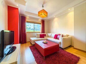 A modern living room with a sectional sofa, a red ottoman, a flat-screen TV, and a large window with curtains. - Refúgio do Mar by ACasaDasCasas (Ericeira)