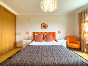 Ein Schlafzimmer mit einem großen Bett, zwei Nachttischen mit Lampen, einem orangefarbenen Sessel, einer Wanddekoration und einer Chandelier.