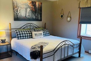 A bedroom with a metal bed frame, a white bedspread, a welcome pillow, a wall-mounted painting, and a window with blinds.
