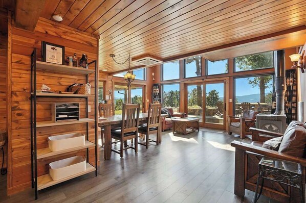 A spacious room with a wooden ceiling, large windows, a dining area with chairs, a sofa, and a fireplace.
