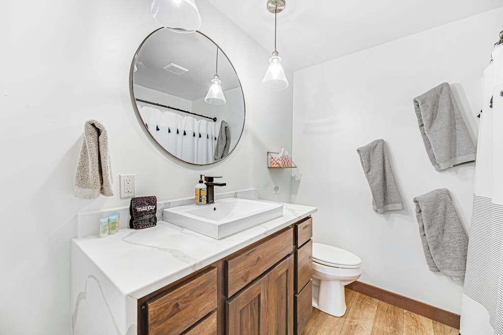 A modern bathroom with a round mirror, white sink, wooden cabinets, and a toilet.