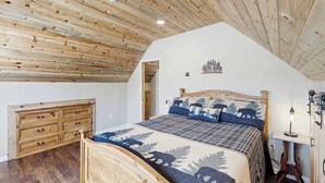 A bedroom with a wooden ceiling, a bed with a forest-themed bedspread, a wooden dresser, and a small bedside table with a lamp. - Five Mile Retreat- Spacious Retreat • Large Deck & WiFi (Cripple Creek)