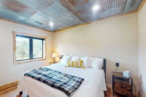 A bedroom with a bed, a window, a bedside table, and a lamp. - Bear Butte Retreat - Private Hot Tub Retreat • Secluded (Deadwood)