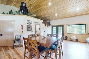 A dining area with a wooden table and chairs, a bear head decoration on the wall, and a window with a view of trees. - Bear Butte Retreat - Private Hot Tub Retreat • Secluded (Deadwood)
