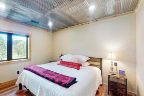 Ein Schlafzimmer mit einem Bett, einem Nachttisch, einer Lampe und einem Fenster mit Blick auf Bäume.