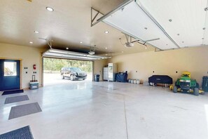 A spacious garage with a car and a green tractor, a blue door, and a ceiling fan. - Bear Butte Retreat - Private Hot Tub Retreat • Secluded (Deadwood)