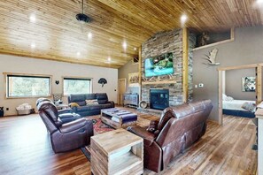 A spacious living room with leather sofas, a stone fireplace, and a television. - Bear Butte Retreat - Private Hot Tub Retreat • Secluded (Deadwood)