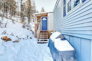 Schneebedecktes Freigelände mit einer Holztreppe, die zu einer blauen Tür führt, umgeben von Bäumen.
