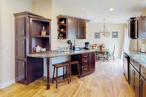 A kitchen with dark wood cabinets, a central island with stools, a chandelier, and a dining area with chairs. - The Great Escape - Dog-Friendly Cabin • Views Near Deadwood (Deadwood)