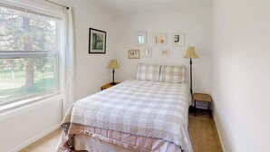 Ein Schlafzimmer mit großem Fenster, einem Bett mit kariertem Überwurf, Nachttischlampen und gerahmten Bildern an der Wand.