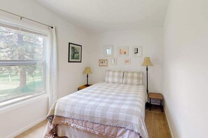 Ein Schlafzimmer mit großem Fenster, einem Bett mit kariertem Überwurf, Nachttischlampen und gerahmten Bildern an der Wand.