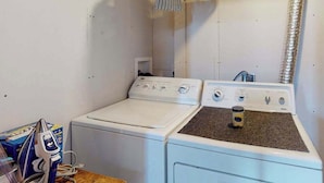 A laundry room with two washing machines and a dryer, a wooden table with a coffee maker, and a vent on the wall. - Springs Sanctuary - Dog-Friendly Home • Decks & Fireplaces (Colorado Springs)